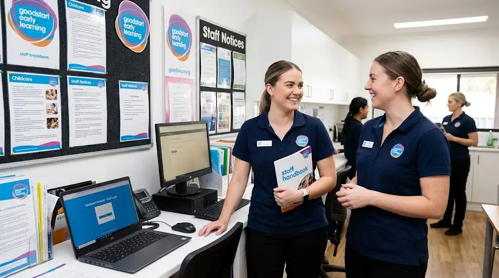Two Goodstart Early Learning employees in uniform reviewing a staff handbook next to a laptop displaying the Goodstart intranet staff login portal.